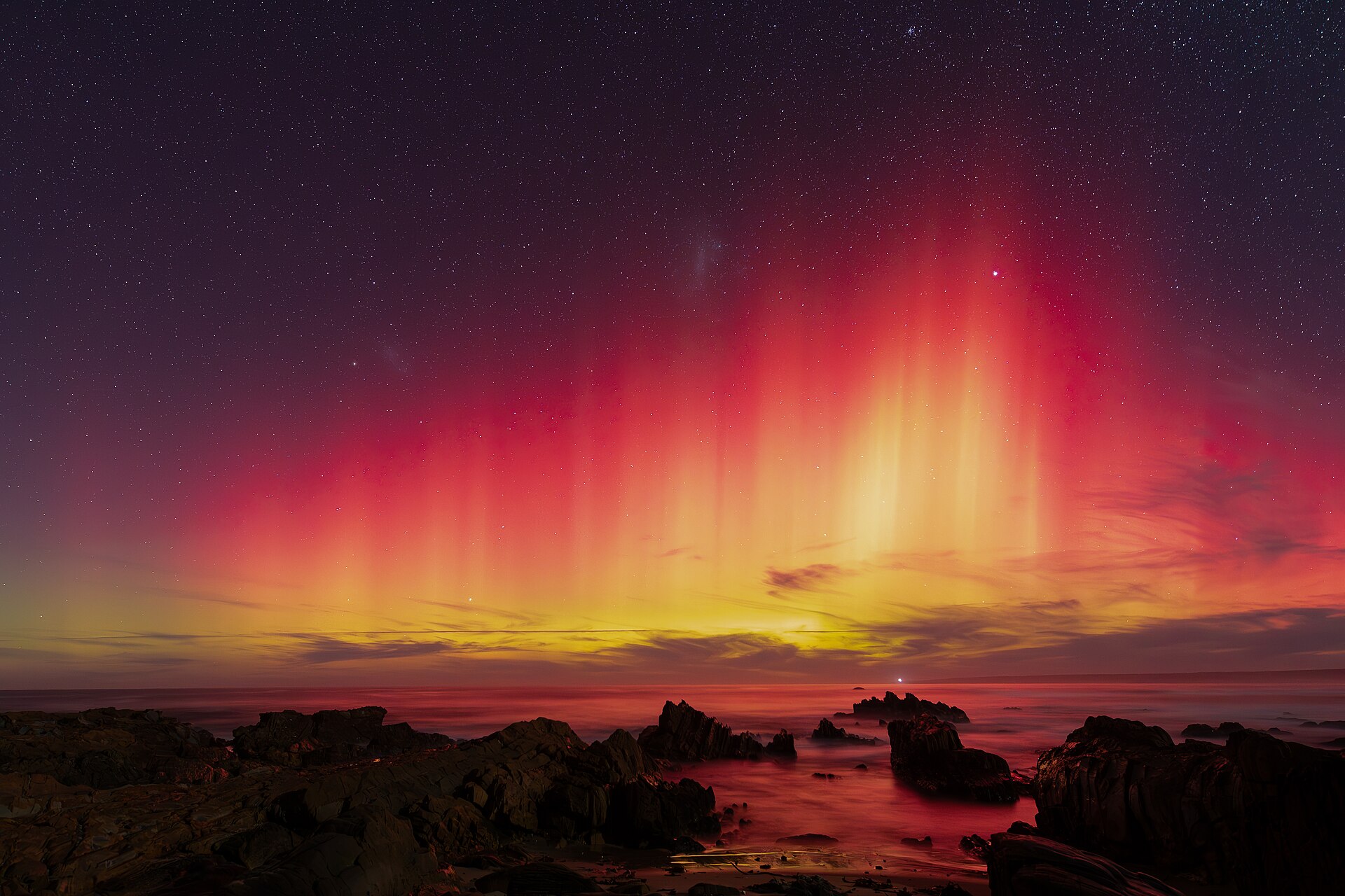 Red and yellow aurora australis rays above rocks and the sea at Mallacoota, Victoria, under a starry night sky.