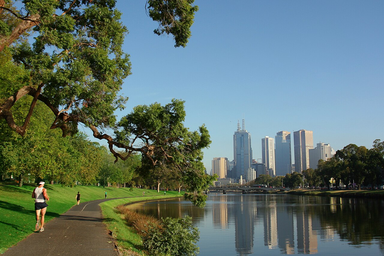 A runner on a path beside a tree-lined river with tall city towers in the distance, reflected in the water under a clear blue sky.