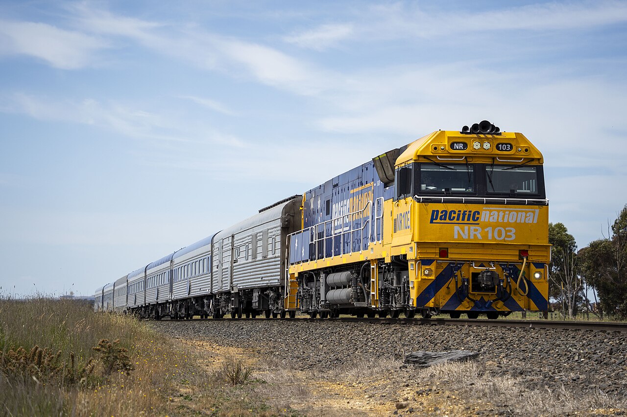A yellow and blue Pacific National diesel locomotive numbered NR 103 pulls a train of silver passenger carriages along a gravel-ballasted track. Dry grass is in the foreground, a few trees stand at the right and pale clouds fill the blue sky.