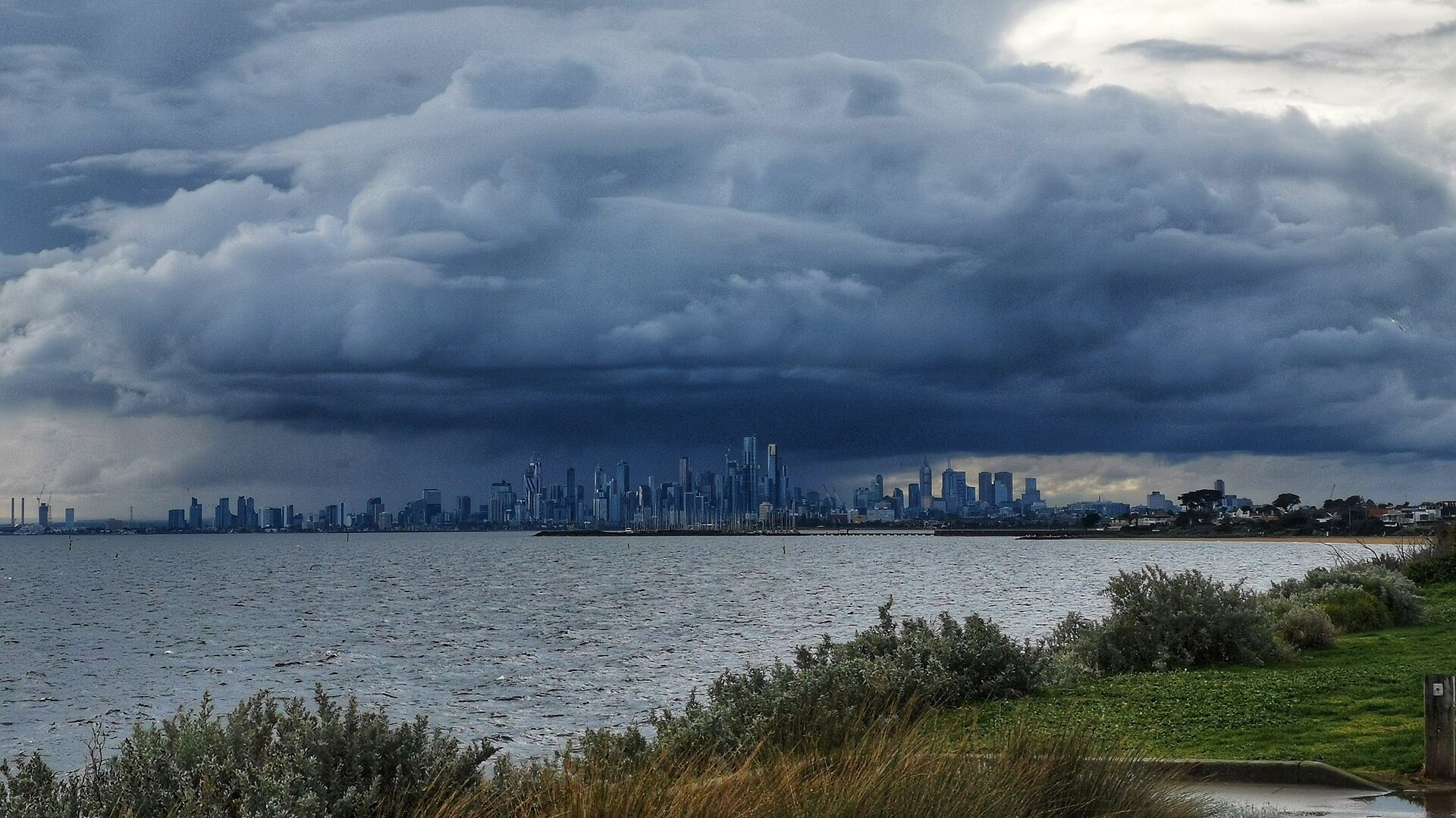 Dark storm clouds above the Melbourne skyline seen across water, with grass and shrubs in the foreground.