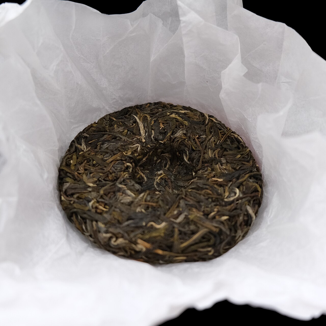 A round pressed disc of dark and olive twisted tea leaves with a shallow dimple in the centre, sitting in crumpled white tissue paper against a black background.