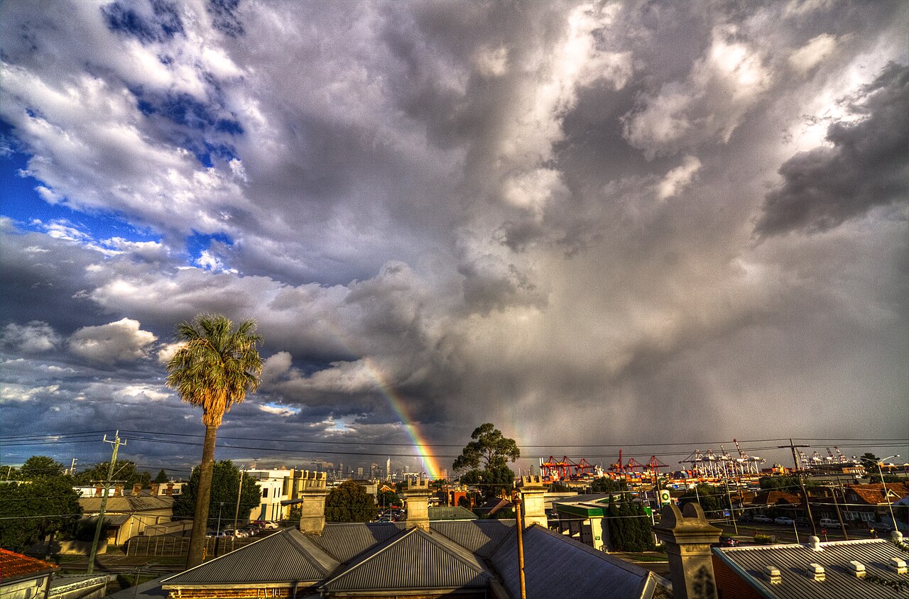 Thick grey and white storm clouds fill the sky over suburban rooftops, with a rainbow reaching down near the distant Melbourne skyline and port cranes on the horizon. A palm tree stands at the left.