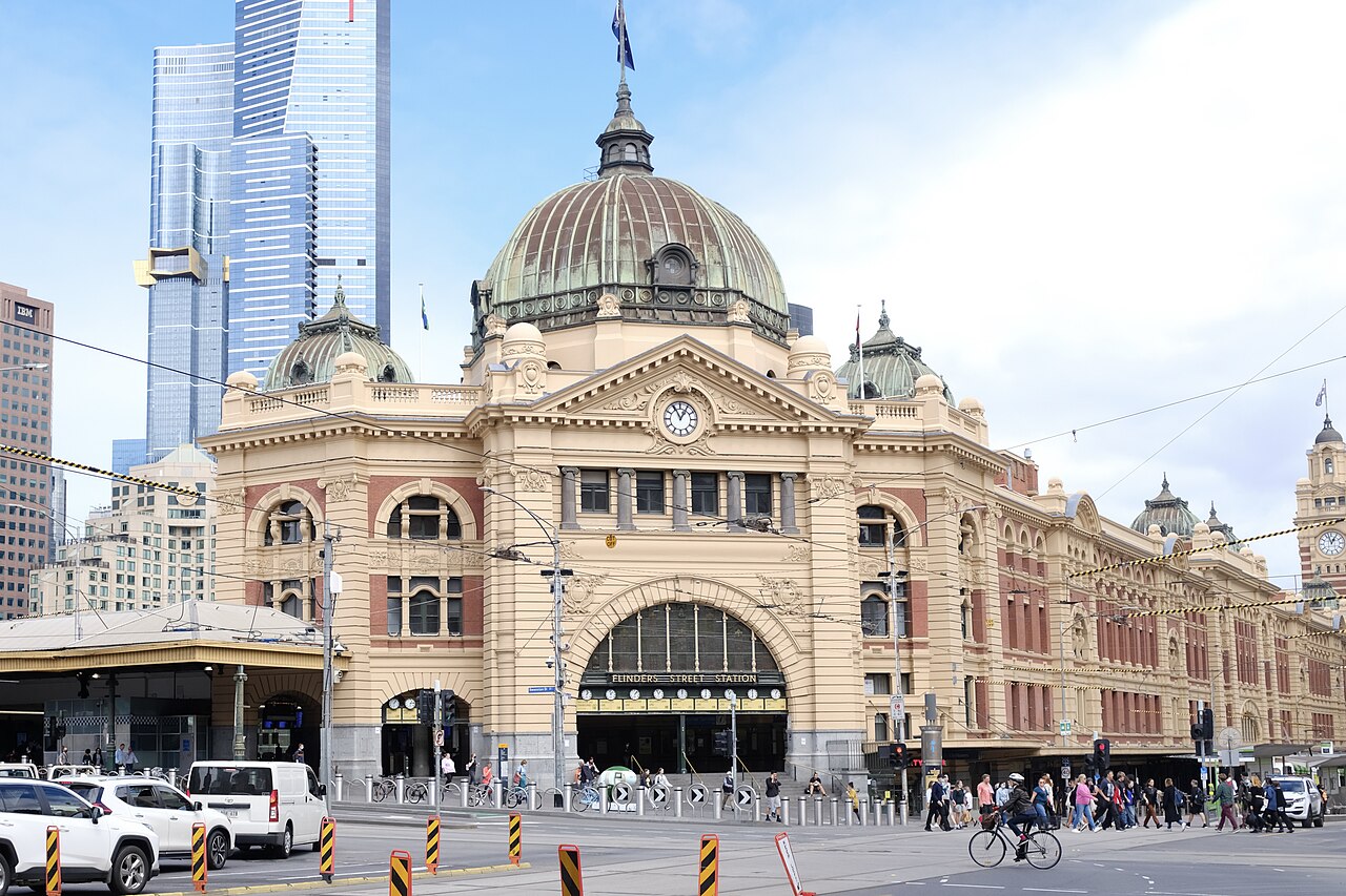 The domed, cream and red-brick front of Flinders Street Station in Melbourne, with tall towers behind it, cars and traffic bollards in the foreground and people crossing the street on the right.